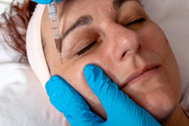 Güzellik uzmanı müşterinin yanağına hyaluronik asit enjekte ederek göz çevresindeki kırışıklıkları kapatıyor. Orta yaşlı bir kadın güzellik salonunda kırışıklık önleyici botoks tedavisi görüyor..