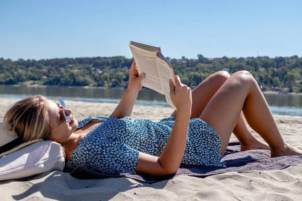 一个戴着太阳镜的年轻貌美的女人正在看书 独自在荒芜的沙滩上消磨时光 — 图库照片