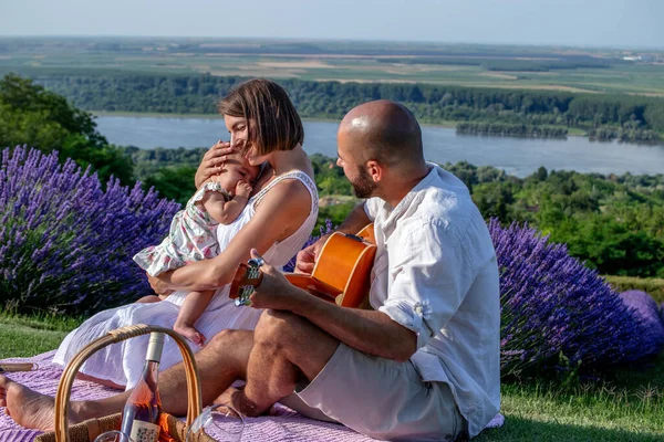 川沿いの美しいラベンダー畑で小さな赤ちゃんの女の子と一緒にピクニックを楽しむ愛の若いカップル 夫は彼の素敵な家族にギターを弾いている — ストック写真