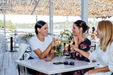 Üç neşeli olgun kız arkadaş bir masada oturmuş kadeh kaldırıyor, sohbet ediyor ve nehir kıyısındaki bir restoranın terasında gülüyor..