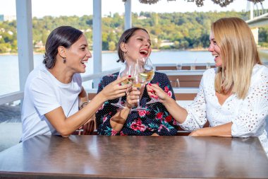 Üç neşeli olgun kız arkadaş bir masada oturmuş kadeh kaldırıyor, sohbet ediyor ve nehir kıyısındaki bir restoranın terasında gülüyor..