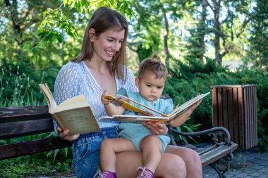 Anne, küçük kızıyla birlikte parkta bir bankta oturuyor ve birlikte kitap okuyor. Çocuk gelişimi ve eğitim.