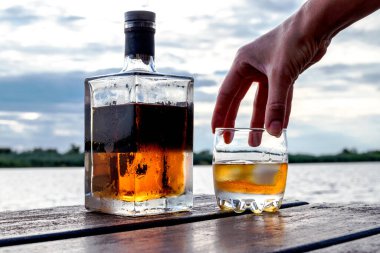 A chilled bottle and a glass of spirits on rocks are on a wooden dock by the water. Heavy clouds at sunset in the background, while the human hand reaches for the glass. clipart