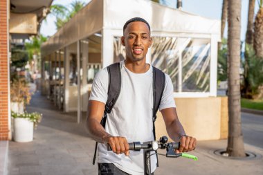 Güneşli bir günde sokakta elektrikli scooterla gezen sıradan giyinmiş mutlu, Afro-Amerikan bir adam. Scooter süren neşeli yakışıklı adam.