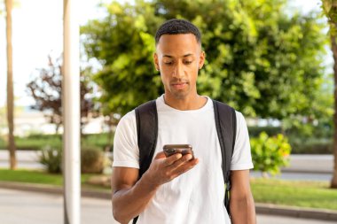 Kentin caddesinde akıllı telefon kullanarak gülümseyen Afro-Amerikalı genç adamın yan görüntüsü.