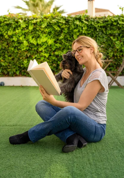 Young woman sitting in a park and reading a book with her dog.