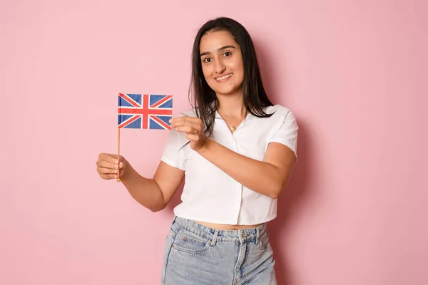 穿着白色休闲装T恤 举着英国国旗 庆祝节日 看着相机 满意地笑着的女性画像 在粉色背景下被隔离的室内拍摄 — 图库照片