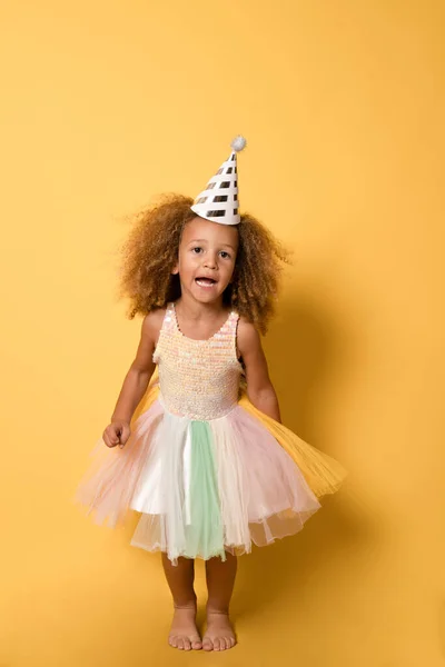 stock image Funny child girl with party hat wearing a festive dress standing isolated over yellow background.