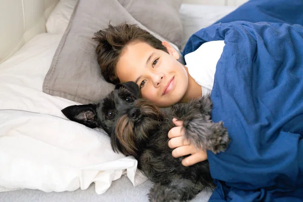 ベッドに横になって犬と一緒にカメラで笑っている幸せな笑顔の子供の男の子 幸せな子供たちの家で楽しい時間を過ごすコンセプト — ストック写真
