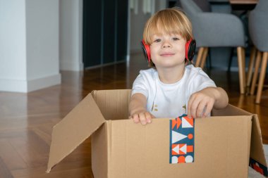 Kulaklıklı küçük çocuk yeni evinde karton bir kutuyla eğleniyor..