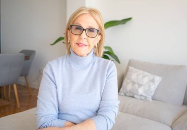 Portrait of beautiful smiling senior woman with blonde hair sitting on the sofa at home.