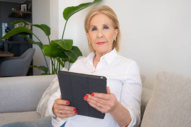 Dijital tablet kullanan, gülümseyen olgun yaşlı bir kadın kanepeye oturur, e-kitap okur, mutlu orta yaşlı büyükanne evde bilgisayarında internet alışverişi yapıyor.