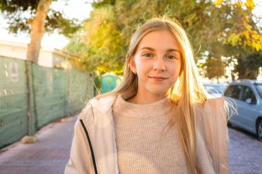 Okuldan sonra sırt çantalı güzel bir kız çocuğu gülümsüyor ve kameraya bakıyor. Sonbaharda açık havada genç bir kızın portresi.