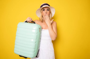 Amazed woman wearing straw hat holding suitcase standing isolated over yellow background.
