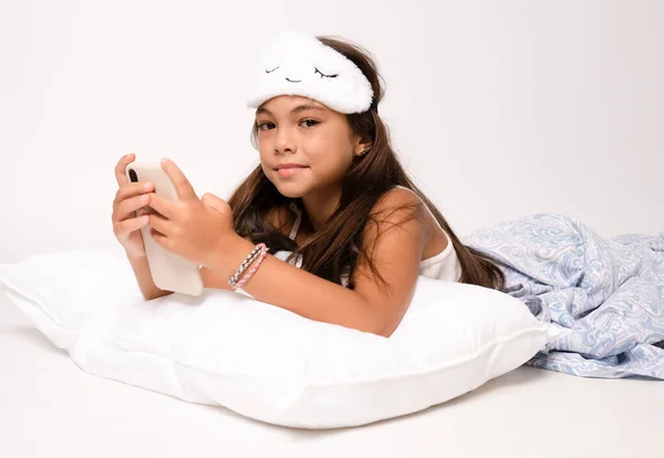 Child girl in sleep wear lying on floor using smartphone isolated over white background.