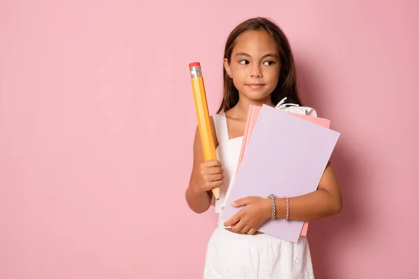 Smiling active excellent best student schoolgirl holding books and a big pencil going to school isolated in pink background