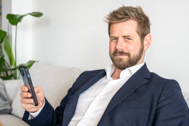 Gülümseyen Kafkasyalı genç adam rahat ol. Evdeki koltuğa otur ve cep telefonu cihazını kullan. Mesaj at. Akıllı telefon cihazında milenyum erkek kablosuz internetin kutlu olsun. İletişim konsepti.