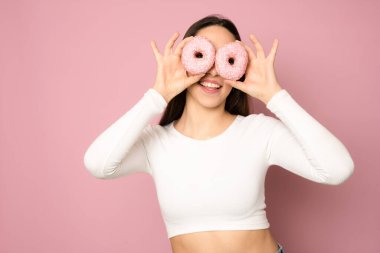 smiling girl having fun with sweets isolated on pink background. Attractive young woman with brown hair in white t-shirt posing with doughnuts in her hands