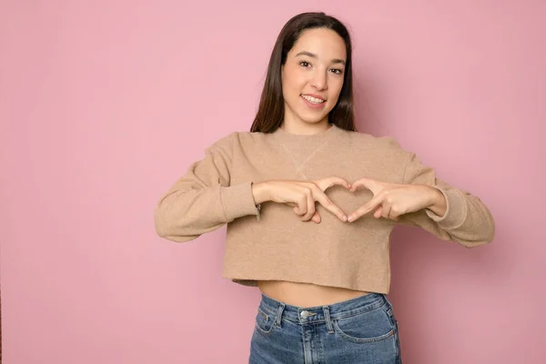 Cute beautiful girl in love making heart with fingers and smiling isolated over pink background.