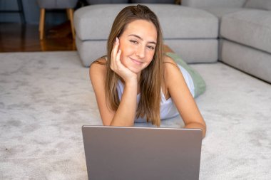 Dizüstü bilgisayar kullanan mutlu genç bir kadın evdeki halıya uzanmış kameraya bakıyor.
