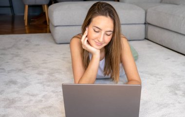 Evdeki halıda dizüstü bilgisayar kullanan mutlu genç kadın.