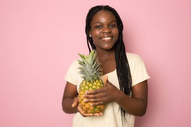 Genç siyahi kadın pembe arka planda izole edilmiş ananasla poz verirken gülüyor.