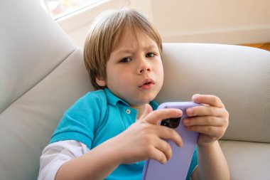 Akıllı telefon kullanan sevimli küçük çocuğu kapat, ekrana bak, elinde telefonu tutan meraklı çocuk, koltukta tek başına oturuyor, mobil cihaz oyunu oynuyor, çevrimiçi çizgi film izliyor..