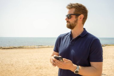 Akıllı telefon kullanan yakışıklı bir portre adamı, mutlu yüz, güneş gözlüğü, sakal, plaj, deniz, seyahat, internet, açık hava portresi, hippi tarzı, telefonu elinde tutan, mesajlara cevap veren. sakallı adam.