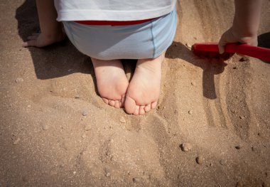 Tatilde mayo giymiş, deniz kenarındaki kumsalda oynayan beyaz, şirin bir çocuğun ayak görüntüsü.