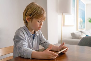 Akıllı telefon kullanan sevimli küçük çocuğu kapat, ekrana bak, elinde telefonu tutan meraklı çocuk, sandalyede tek başına oturuyor, mobil cihaz oyunu oynuyor, internetten çizgi film izliyor, sohbet ediyor.