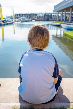 Panamalı küçük bir çocuk deniz manzaralı banktan denize açılan yatı seyrediyor. Küçük çocuk şehir seti üzerinde dinleniyor.
