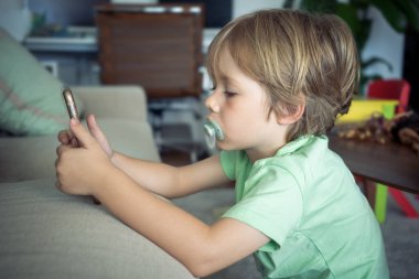 Akıllı telefon kullanan sevimli küçük çocuğu kapat, ekrana bak, elinde telefonu tutan meraklı çocuk, koltukta tek başına oturuyor, mobil cihaz oyunu oynuyor, çevrimiçi çizgi film izliyor..