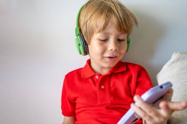 Kablosuz kulaklık takan ve cep telefonu kullanan bir anaokulu öğrencisi gülümsüyor. Video sohbeti, cep telefonu, çocuklar ve teknoloji kavramı.