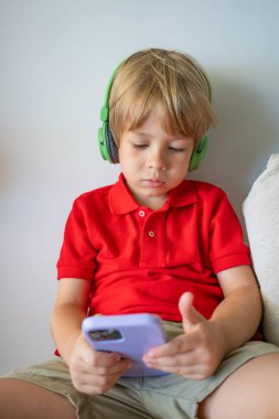 Kablosuz kulaklık takan ve cep telefonu kullanan bir anaokulu öğrencisi gülümsüyor. Video sohbeti, cep telefonu, çocuklar ve teknoloji kavramı.