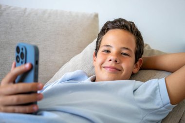 Evdeki kanepede uzanmış akıllı telefon kullanan rahat bir çocuk.