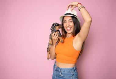 Hasır şapka takan ve pembe arka planda izole edilmiş dachshund köpeği kucaklayan tatlı esmer kadın. Hayvanlara sevgi, evcil hayvan konsepti. Dachshund köpeğini tutan neşeli kadın.