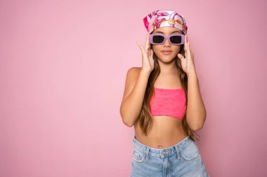 Yaz kıyafetleri içinde güneş gözlüğü takan sevimli bir kızın portresi pembe arka planda tek başına duruyor..