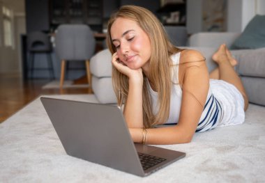 Evde dizüstü bilgisayar kullanarak yerde yatan neşeli genç bir kadın görüntüsü.