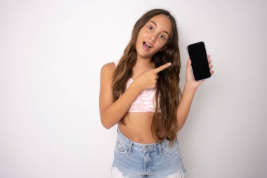 Beyaz duvara karşı yaz kıyafetleri giyen heyecanlı beyaz kız beyaz ekranlı akıllı telefonu işaret ediyor. Reklam kavramı.