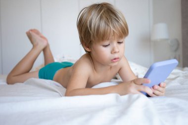 Evde yatakta uzanmış akıllı bir telefonla oyun oynayan gülümseyen bir çocuğun portresi. Çocuk bir otel odasında cep telefonu kullanıyor. Kumar bağımlılığı kavramı. Eğlenen bir çocuk. Gadget ellerde.