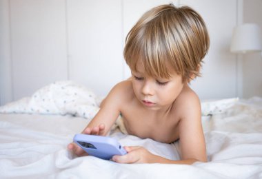 Evde yatakta uzanmış akıllı bir telefonla oyun oynayan gülümseyen bir çocuğun portresi. Çocuk bir otel odasında cep telefonu kullanıyor. Kumar bağımlılığı kavramı. Eğlenen bir çocuk. Gadget ellerde.