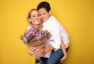 Oğlu mutlu annesine çiçeklerle sarılıyor. Anneler günü konsepti. Aile tatili. Sarı arkaplanda izole edilmiş