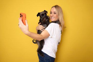 Arkadaşlık kavramı. Şirin köpeğiyle selfie çeken mutlu Avrupalı kadın sarı stüdyo arka plan duvarının üzerinden kameraya bakıp gülümsüyor.