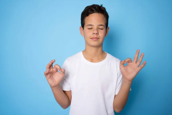 Handsome Caucasian teenager boy wearing white t-shirt relax and smiling with eyes closed doing meditation gesture with fingers. Yoga concept.