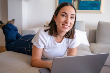 Güzel genç bir kadın evinde kanepede otururken dizüstü bilgisayar kullanıyor ve gülümsüyor.