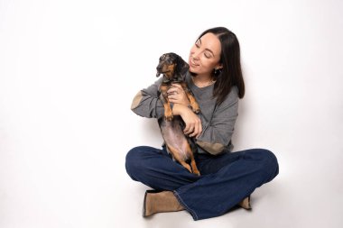 Gülümseyen genç bir kadın yerde köpeğinin yanında tek başına beyazların üzerinde oturuyor.