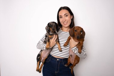 Sıradan çizgili tişört giyen genç bir kadın beyaz arka planda soyutlanmış iki köpeğine sarılıyor..