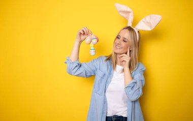 Gülümseyen neşeli, neşeli, sıradan kıyafetler giyen genç bir kadın tavşan kulakları sepet sepetinde sarı arka plan stüdyo resminde izole edilmiş renkli yumurtalar tutuyor. Mutlu Paskalyalar