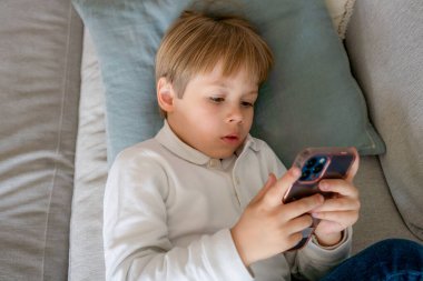 Kanepede uzanmış akıllı telefon kullanan şirin bir çocuk.