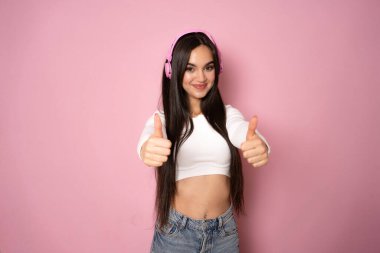 Genç mutlu, eğlenceli Latin kadın gündelik kıyafetler giyer ve kulaklık takar. Pembe arka plan stüdyo resminde baş parmaklarını göstererek müzik dinler. Yaşam tarzı kavramı